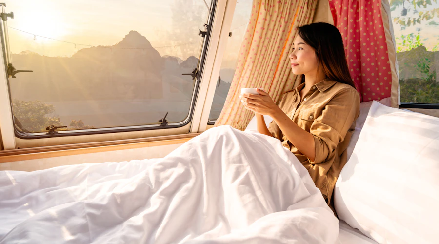 A woman drinks coffee in her camper bed with white sheets